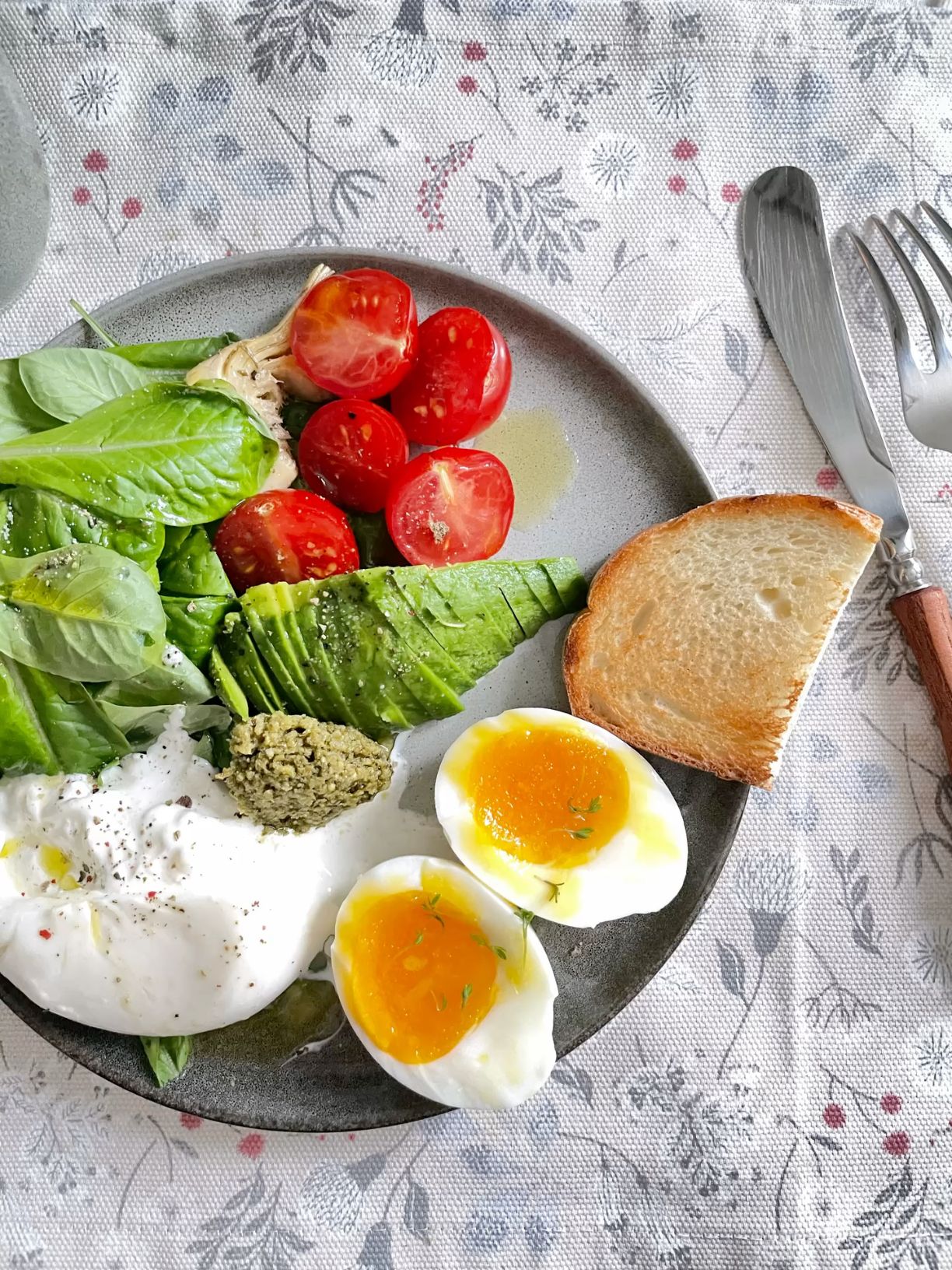 Breakfast plate with soft-boiled eggs, sliced avocado, burrata cheese, cherry tomatoes, salad, and toast on a patterned tablecloth